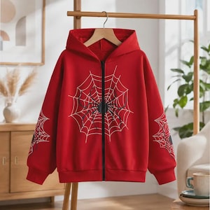 May include: A bright red zip-up hoodie featuring a large white spiderweb graphic on the front and smaller spiderweb designs on each sleeve. The garment includes a hood, long sleeves with ribbed cuffs, and a black vertical zip closure down the centre.