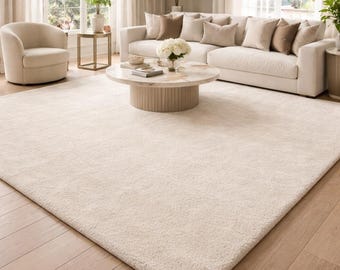 Puede incluir: Una gran alfombra rectangular de color crema domina el primer plano de un salón. La alfombra tiene una superficie suave y texturizada. También se ven un sofá seccional, un sillón y una mesa de centro, creando un ambiente acogedor.