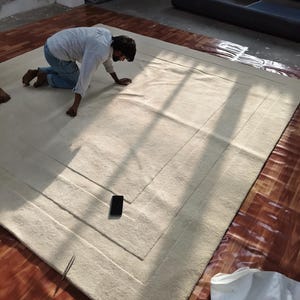 May include: A large, rectangular, off-white rug with a bordered design. The rug is on a wooden floor. A person is kneeling on the rug, inspecting it. A black smartphone and a pair of tweezers are on the rug.