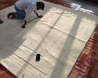 May include: A large, rectangular, off-white rug with a bordered design. The rug is on a wooden floor. A person is kneeling on the rug, inspecting it. A black smartphone and a pair of tweezers are on the rug.