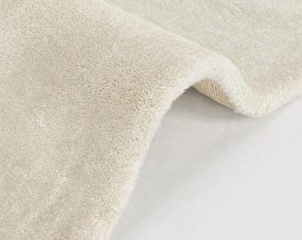 Peut inclure: Gros plan sur un tapis crème avec une surface douce et texturée. Le tapis est partiellement enroulé, révélant le dessous. La couleur neutre et la texture moelleuse suggèrent un élément de décoration intérieure confortable.