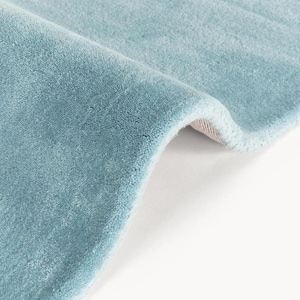 May include: A soft, light blue area rug with a dense, plush pile texture. The rug is folded over to reveal a glimpse of the light-colored backing material underneath, showcasing the thickness and quality of the fabric construction.