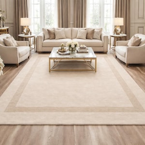 Può includere: Un grande tappeto beige rettangolare con un bordo beige più chiaro e un bordo interno beige più scuro. Il tappeto si trova in un soggiorno con un divano, sedie e un tavolino in marmo. La stanza ha toni neutri e luce naturale.