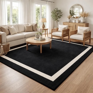 May include: A large, rectangular black area rug with a wide, off-white border. The rug is placed in a living room setting with a sofa, chairs, and a coffee table. The rug's dimensions are approximately 8 x 10 feet.