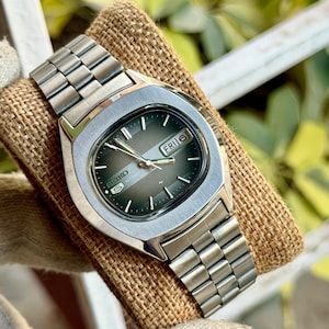 May include: A Seiko 5 automatic wristwatch with a silver-tone stainless steel link bracelet and a cushion-shaped case. The watch features a dark green gradient dial with silver baton indices, silver hands, and a day-date window displaying FRI 6.