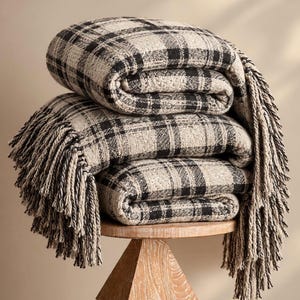 May include: A folded throw blanket featuring a black and beige plaid pattern with a textured weave. The blanket has long, thick fringe detailing along the edges, creating a soft and layered appearance as it sits stacked in multiple folds.