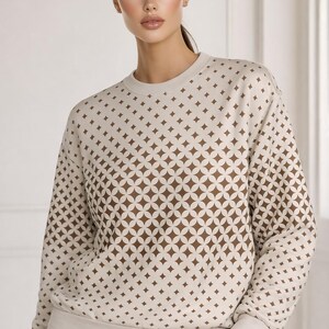 Peut inclure: Un sweat-shirt à manches longues et col rond de couleur beige clair, orné d'un motif géométrique marron répétitif composé de cercles entrelacés et d'étoiles à quatre branches. Le vêtement présente un col, des poignets et un ourlet côtelés.