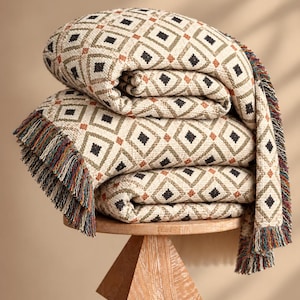 May include: A folded woven throw blanket featuring a repeating diamond pattern in cream, olive green, black, and rust orange. The edges of the blanket are finished with a multi-colored fringe containing threads of blue, red, green, and yellow.