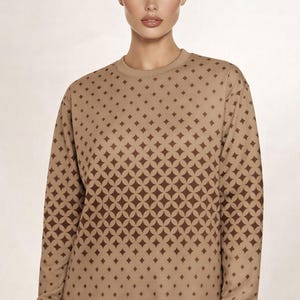 Pode incluir: Uma camisola de manga comprida bege com um padrão geométrico repetitivo de círculos e pequenos losangos castanho-escuros. A peça tem um decote redondo e punhos canelados nas mangas e na bainha para um ajuste estruturado ao corpo.