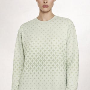 Peut inclure: Un sweat-shirt à manches longues vert sauge clair orné d'un motif géométrique répétitif composé de cercles entrelacés et d'étoiles à quatre branches dans une teinte de vert légèrement plus foncée, avec un col rond et des bords-côtes.