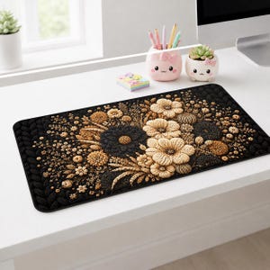 May include: A rectangular desk mat featuring a black background with a textured, knitted-style border and a central floral arrangement in shades of beige, tan, and dark brown. The design displays various flower shapes and wheat-like stalks.