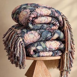 May include: A folded woven throw blanket featuring a dark blue background with a repeating pattern of light pink and lavender protea flowers and green leaves. The blanket has thick, multi-colored twisted fringe along the edges.