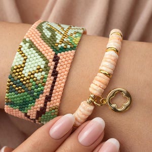 May include: Two bracelets are displayed. One is a wide, beaded bracelet with a geometric pattern in green, peach, and brown. The other is a strand of peach-coloured beads with gold accents and a gold charm.