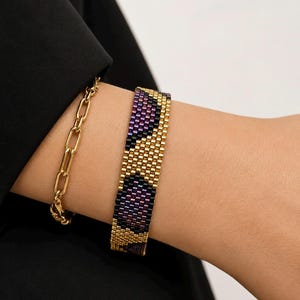 Könnte beinhalten: Ein breites gewebtes Perlenarmband mit einem geometrischen Muster aus metallisch goldenen, dunkelvioletten und schwarzen Glasperlen. Daneben befindet sich ein goldfarbenes Gliederarmband mit länglichen ovalen Gliedern, beide zusammen getragen.