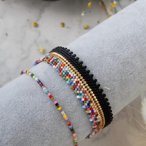 Könnte beinhalten: Drei Armbänder mit Perlen. Das obere Armband ist schwarz mit goldenen Akzenten und bunten Perlen. Das zweite Armband hat ein goldenes Band mit bunten Perlen. Das dritte Armband ist ein dünner Strang aus bunten Perlen.