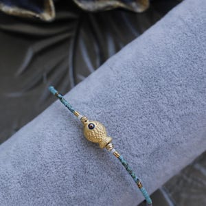 Könnte beinhalten: Armband mit goldenem Fischanhänger und türkisfarbenen Perlen. Der Fischanhänger hat ein dunkelblaues Auge und ist in der Mitte des Armbands. Das Armband besteht aus kleinen türkisfarbenen Perlen und goldfarbenen Akzenten.