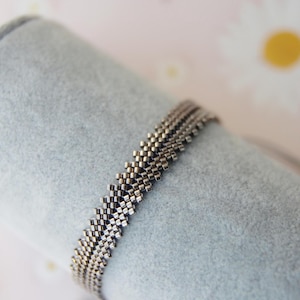 May include: A silver beaded bracelet with a geometric pattern. The bracelet is made of small, square beads and is displayed on a light blue surface. The background has a floral pattern with white flowers.