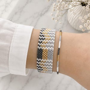 Könnte beinhalten: Ein breites Perlenarmband und ein schmales Perlenarmband mit einem geometrischen Muster in Anthrazit, Hellgrau, Weiß und Metallic-Gold. Die Armbänder bestehen aus kleinen, gleichmäßigen, quadratischen Perlen, die in Reihen angeordnet sind.