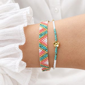 Op de afbeelding: Twee armbanden die samen worden gedragen. De ene is een brede, platte geweven band met een diagonaal chevronpatroon van roze, turquoise, witte en gouden kralen. De tweede is een dun, delicaat strengetje van witte en turquoise kralen met een kleine gouden bloembedel.