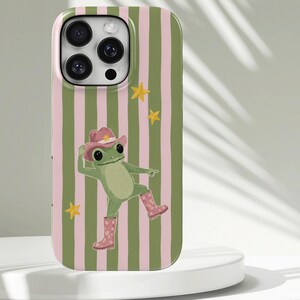 Puede incluir: Una funda para smartphone con un patrón de rayas verticales en verde oliva y rosa claro. El diseño incluye una ilustración central de una rana verde que lleva un sombrero de vaquero rosa y botas de lluvia rosas a juego, rodeada de estrellas amarillas.
