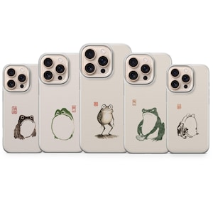 May include: Five light-colored smartphone cases arranged in a row, each featuring a unique ink-style illustration of a frog and a small red square stamp. The designs vary from a seated frog to a standing frog and a frog holding a small bowl.
