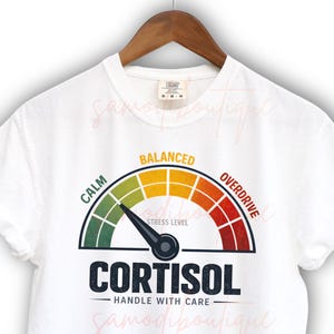 Könnte beinhalten: Weißes T-Shirt mit einer Grafik eines Stresspegelmessers. Das Messgerät zeigt "Cortisol" an und hat eine Nadel, die auf die Zone "Calm" zeigt. Das Shirt enthält auch den Text "Handle with Care."
