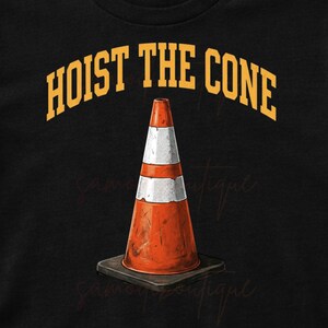May include: Black t-shirt with the text "HOIST THE CONE" in yellow arched lettering above a graphic of an orange and white traffic cone. The cone is on a black square base. The shirt is displayed with jeans, sunglasses, and white sneakers.