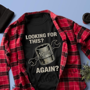 Puede incluir: Camiseta negra con el texto "LOOKING FOR THIS?" y un gráfico de un vaso de 10 mm y una llave. También se ven una camisa a cuadros roja y negra, un gorro negro, gafas de sol y una planta pequeña.