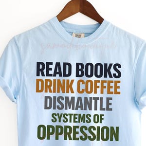 Puede incluir: Camiseta azul claro con el texto "READ BOOKS DRINK COFFEE DISMANTLE SYSTEMS OF OPPRESSION" en negro, marrón, gris y verde. La camiseta está colgada en una percha de madera sobre un fondo blanco. Diseño digital para uso comercial.