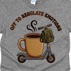 May include: Gray t-shirt with a graphic of a coffee cup on a scooter, a backpack, and the text "OFF TO REGULATE EMOTIONS" in brown. The coffee cup has steam rising from it. The design is centered on the shirt.