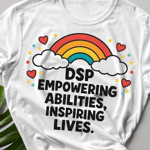 May include: White t-shirt with a rainbow, clouds, and hearts graphic. The shirt has the text "DSP Empowering Abilities, Inspiring Lives." Sunglasses and a black smartphone are next to the shirt. A large green leaf is in the lower left corner.