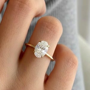 Puede incluir: Un anillo de oro con un diamante de talla ovalada. El anillo tiene una fina banda de oro. El diamante es claro y brillante. El anillo está en un dedo.
