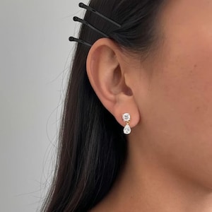 May include: A close-up of a gold-colored earring with a square-cut clear stone and a teardrop-shaped clear stone. The earring is worn on the ear of a person with dark hair and black bobby pins.