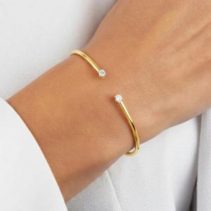 Puede incluir: Un brazalete abierto de color dorado con una pequeña gema redonda y transparente en cada extremo. El brazalete se lleva en una muñeca y el fondo es de tela blanca.