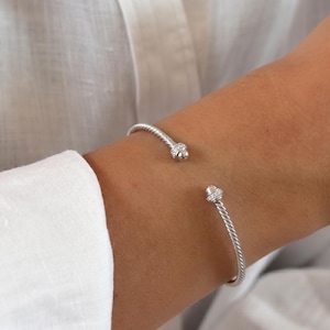 Op de afbeelding: Een zilveren manchetarmband met een gedraaid kabelontwerp. De open armband eindigt in twee afgeronde doppen, elk geaccentueerd met een kleine groep heldere, sprankelende stenen die in het metaal aan de uiteinden van de gedraaide band zijn gezet.
