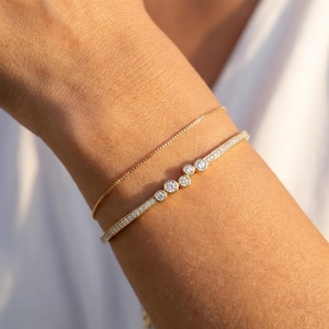 May include: Two gold bracelets are shown on an arm. One is a fine chain, and the other is a gold bangle with small, sparkling stones. The bangle has a curved design with larger stones in the center, creating a stylish look.