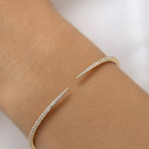 Puede incluir: Una pulsera de puño abierta de color dorado con una fila de pequeñas piedras preciosas transparentes. La pulsera está en una muñeca y los extremos de la pulsera son puntiagudos.