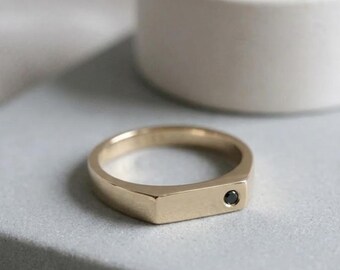 May include: A gold-colored ring with a rectangular face and a small black gemstone. The ring is set on a light gray surface, with a white cylindrical object in the background. The ring is a simple, elegant design.