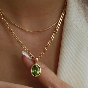 Op de afbeelding: Goudkleurige ketting met een groene ovale hanger. De hanger is gezet in een gouden bezel en hangt aan een gourmetketting. Een tweede, dunnere gouden ketting bevindt zich boven de hangerketting.