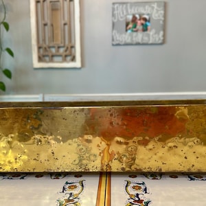 May include: A rectangular brass planter with a hammered, textured finish and a polished gold-tone surface. The container features a long, low-profile design and rests on small, curved metal feet at each corner, showcasing a weathered patina.