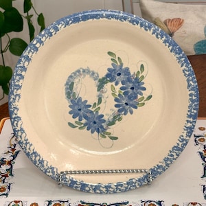 May include: Cream-colored ceramic plate with a blue floral heart design. The plate has a blue speckled border. The heart is made of blue flowers and green leaves. The plate is on a metal stand.