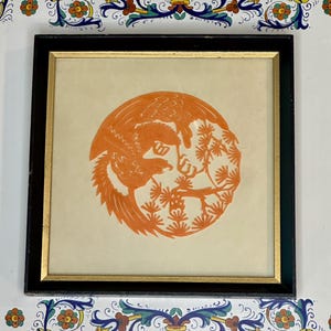 May include: A square framed artwork featuring a circular orange paper cutout of a bird perched on a pine tree branch. The frame is black with a thin gold inner border, set against a white background with blue, green, and orange floral motifs.