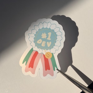 Może przedstawiać: Naklejka w kształcie wstążki z napisem "#1 DEV" w jasnoniebieskim okręgu. Wstążka ma różowe i zielone wstążki oraz małe złote kółko z uśmiechniętą buzią. Naklejka znajduje się na białej powierzchni.
