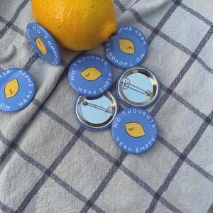 May include: Several blue pin badges, each featuring a yellow lemon illustration and the text "NO THOUGHTS HEAD EMPTY". A fresh lemon sits on a checkered cloth, complementing the badges.