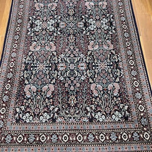 May include: A rectangular, dark blue rug with an intricate floral pattern in shades of red, pink, and white. The rug has a detailed border and fringe along the edges, showcasing a traditional design. The rug is placed on a wooden floor.