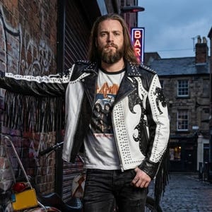 May include: A black and white leather jacket featuring long fringe on the sleeves, silver stud embellishments, and a notched lapel. The jacket is worn over a white t-shirt with a graphic print and is paired with dark denim jeans.