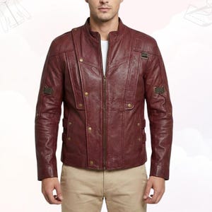 May include: A burgundy leather jacket with a stand-up collar, a front zip, and decorative buttons. The jacket has a textured appearance and features shoulder accents and small rectangular patches. The model is wearing a white shirt underneath.