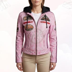 Puede incluir: Chaqueta de moto de cuero rosa con capucha negra y cremallera. La chaqueta presenta múltiples parches con texto y diseños, incluyendo "Limited Edition" y "Race Motorcycles". Rayas moradas verticales recorren las mangas.