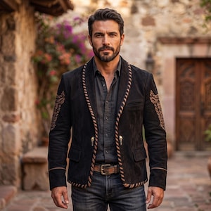 Puede incluir: Un blazer de ante negro con cuello chal y detalles de costuras en marrón, bordados intrincados en color tostado en las mangas y dos botones metálicos con forma de calavera. El blazer se lleva sobre una camisa gris oscuro y vaqueros azules.
