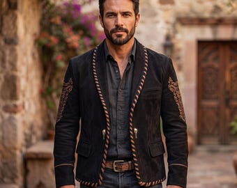 Pode incluir: Um blazer de camurça preta com gola xale e detalhes de costura em castanho, bordados intrincados em tom bege nas mangas e dois botões metálicos em forma de caveira. O blazer é usado sobre uma camisa cinzenta escura e calças de ganga azuis.